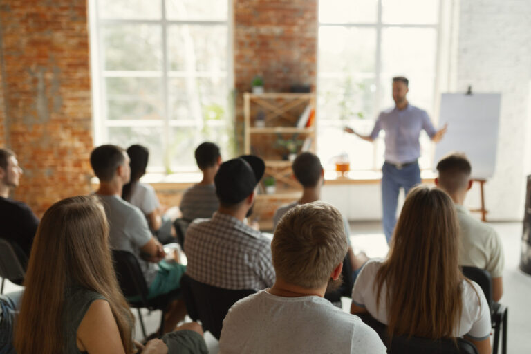 Male speaker giving presentation in hall at university workshop. Audience or conference hall. Rear view of unrecognized participants in audience. Scientific conference event, training. Education concept.